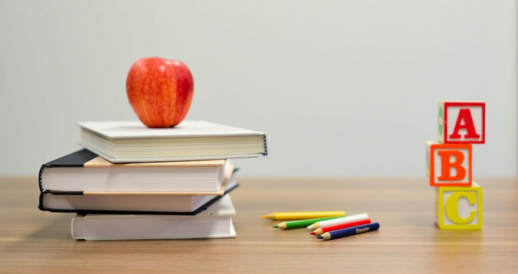 An apple balances on top of a stack of books in a classroom. Also on the desk are pencils and abc blocks.