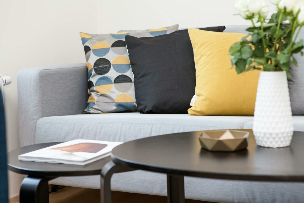 A living room with a coffee table and a grey sofa with grey, black and yellow cushions.